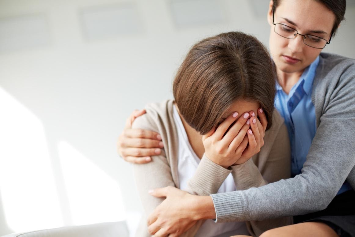 Seated woman with head in hands being comforted in embrace by other woman - guilt and shame can lead to depression